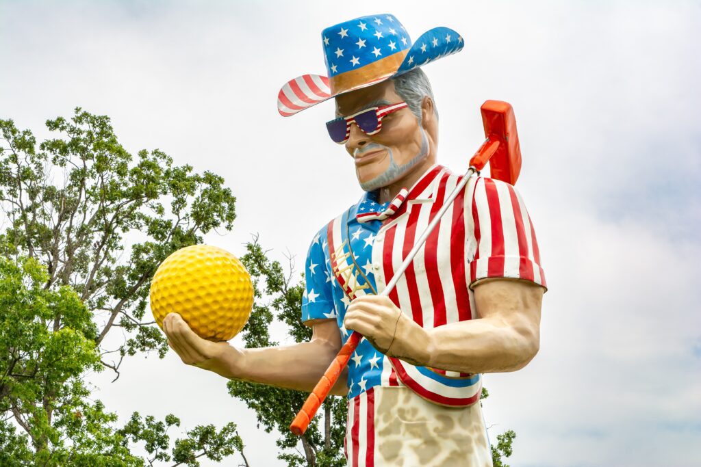 A giant statue of a man dressed in a red, white, and blue patriotic outfit and cowboy hat, holding a large yellow golf ball and an orange golf club, with trees and sky in the background.
