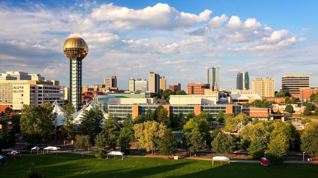 A cityscape of Knoxville, Tennessee, featuring the Sunsphere, a tall structure with a golden glass sphere, surrounded by modern buildings, green trees, and a blue sky with scattered clouds.