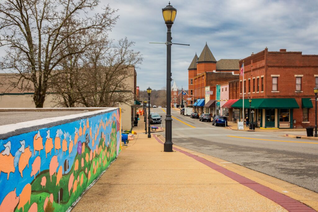 A small-town street with brick buildings, parked cars, and colorful mural on a wall to the left. A lamppost stands on the sidewalk under a cloudy sky.
