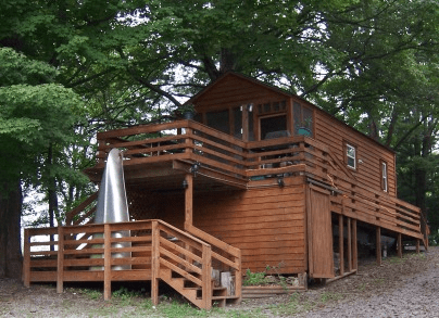 A wooden cabin with a wraparound deck is nestled among trees. The cabin is elevated, has a slide from the deck to the ground, and features multiple windows and a sloped roof.