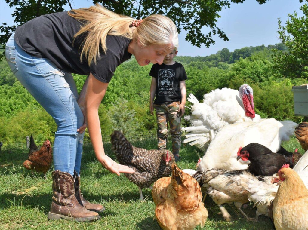 A woman with long blonde hair, wearing jeans and boots, bends down to interact with chickens and turkeys. A child stands behind her in a grassy outdoor area with green trees and blue sky in the background.