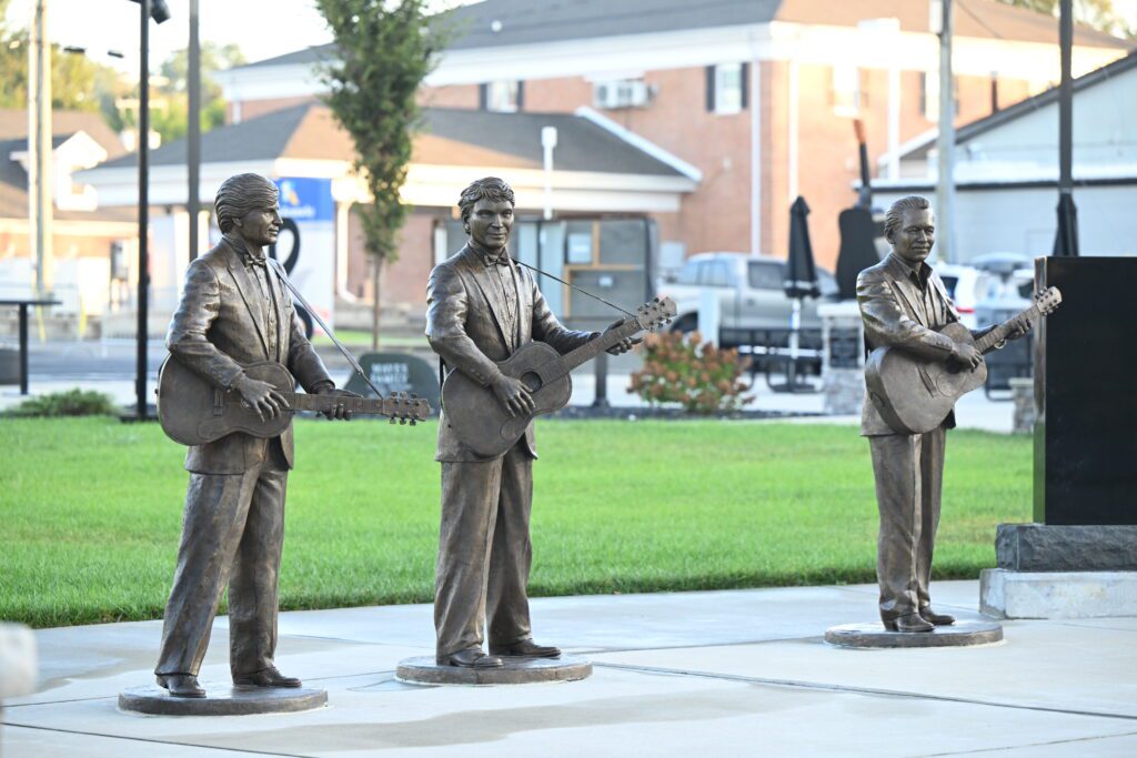 Three bronze statues of men holding guitars stand on a paved area with grass and buildings in the background. The statues are life-sized and arranged in a line, each appearing to play the guitar.