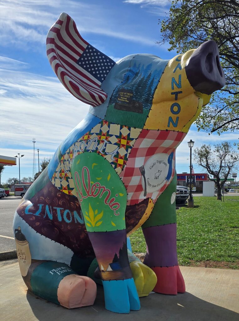 A large, colorful pig statue painted with various patterns, images, and words, including a patchwork quilt, nature scenes, the American flag, and local names, sits on a sidewalk near a grassy area and parking lot.