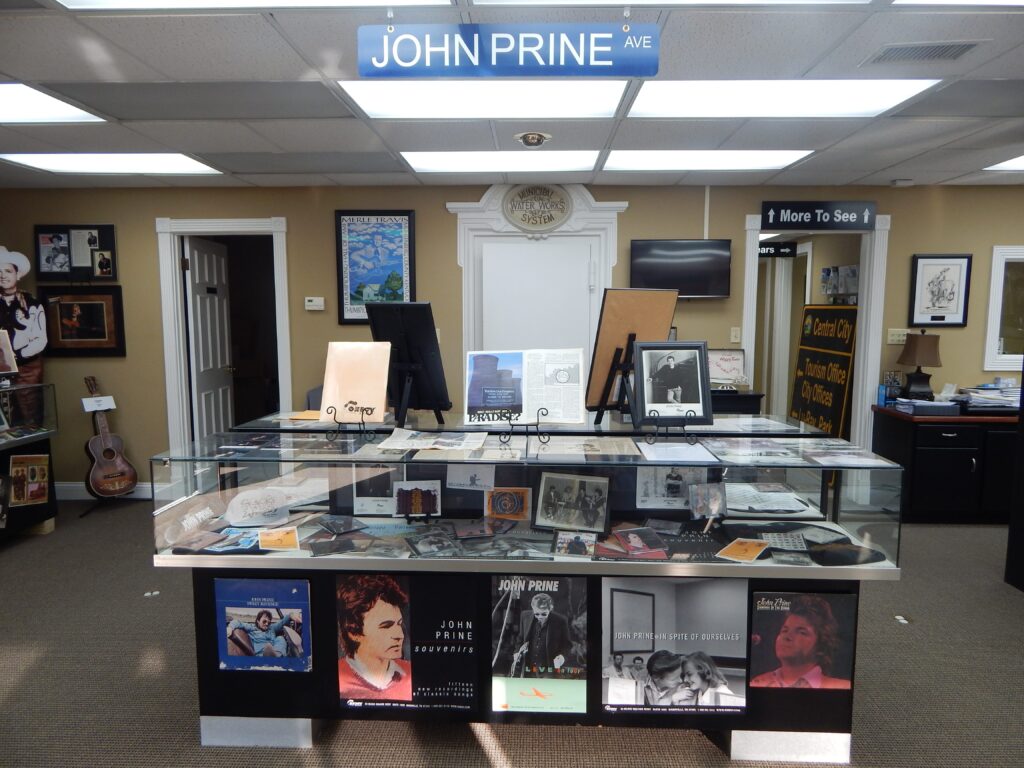 Interior of a music store or museum display dedicated to John Prine, featuring albums, photos, memorabilia in a glass case, and a blue John Prine Ave sign hanging above the display.