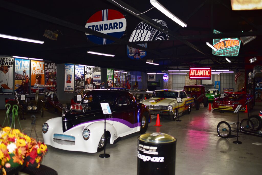 A collection of classic and vintage race cars displayed indoors, surrounded by retro signs, posters, and memorabilia. The cars are brightly colored and arranged in a spacious, well-lit garage or museum setting.
