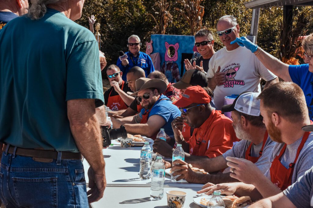 A group of people, some wearing hats and sunglasses, sit at an outdoor table participating in a food-eating contest while others stand around clapping and cheering under sunny weather. Bottles of water and food are on the table.