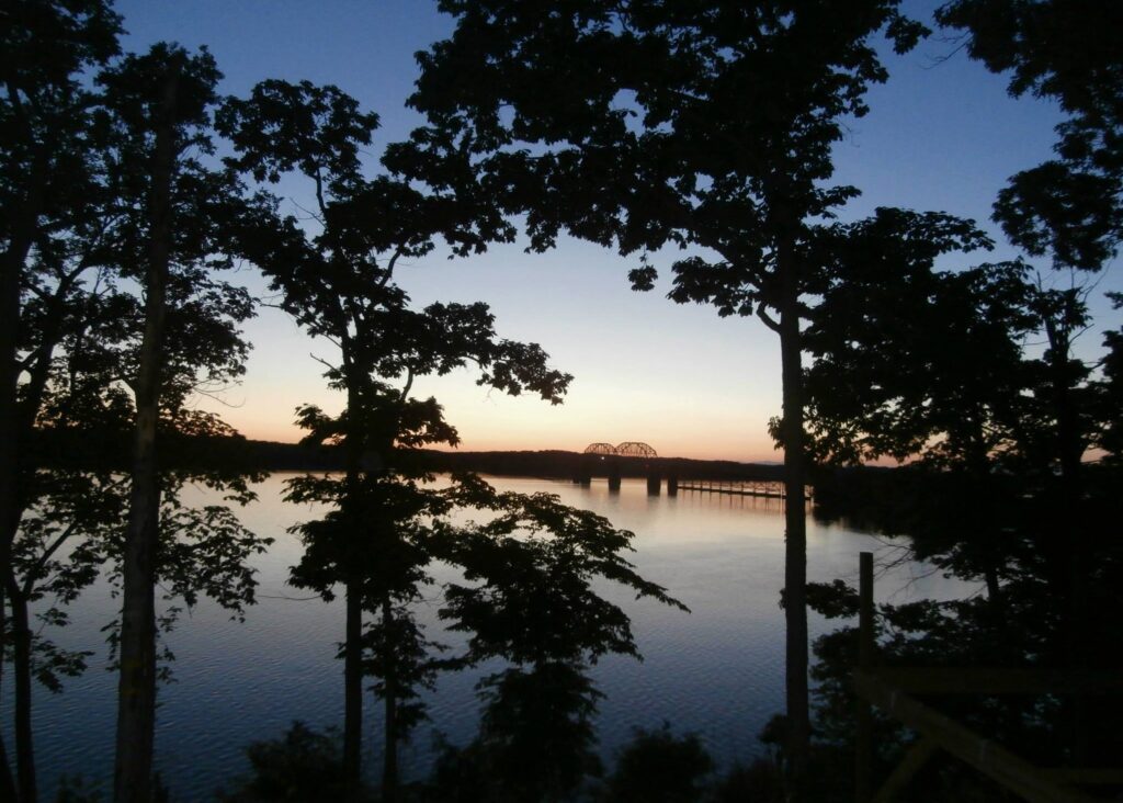 A calm lake at sunset with silhouetted trees in the foreground and a bridge visible in the distance against a fading blue and orange sky.
