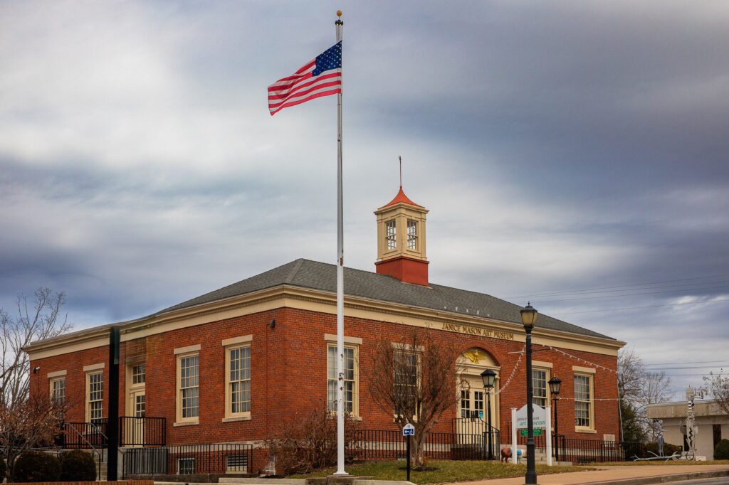 A red brick public building with white trim and a small central clock tower, an American flag on a pole in front, and a cloudy sky overhead. Trees and a lamp post are near the entrance.