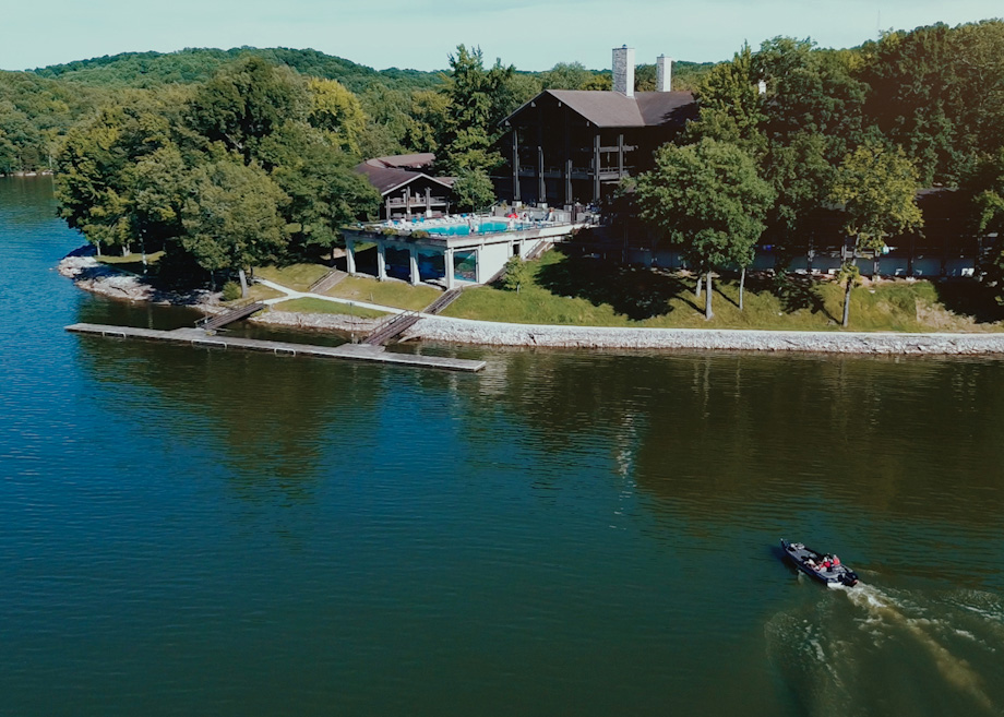 A lodge surrounded by trees sits on the edge of a lake. A pool with people is visible on a terrace, and a small motorboat moves across the water in the foreground.