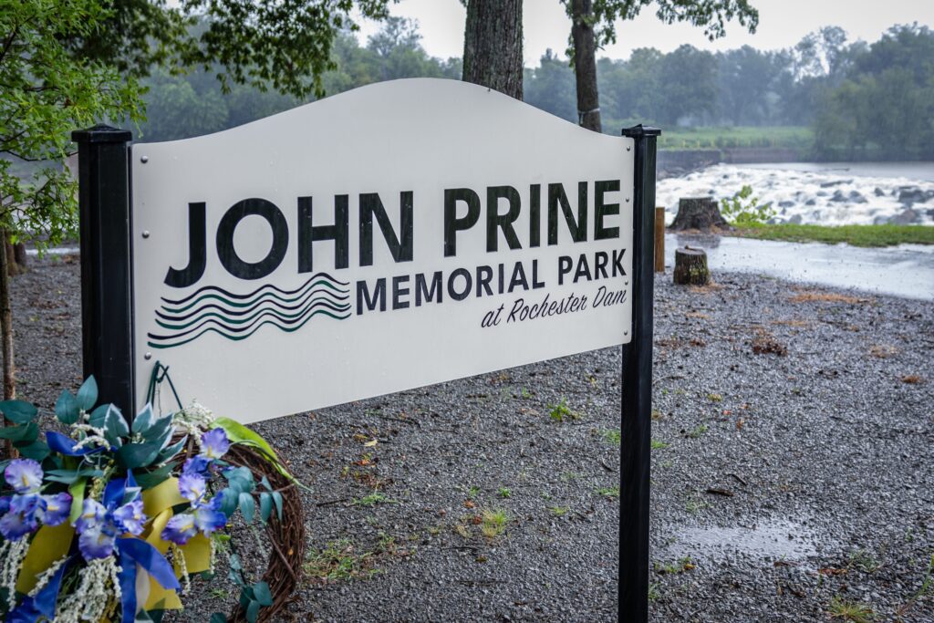A white sign reads “John Prine Memorial Park at Rochester Dam,” standing near trees and a riverbank on a cloudy day, with a blue and yellow floral wreath nearby.