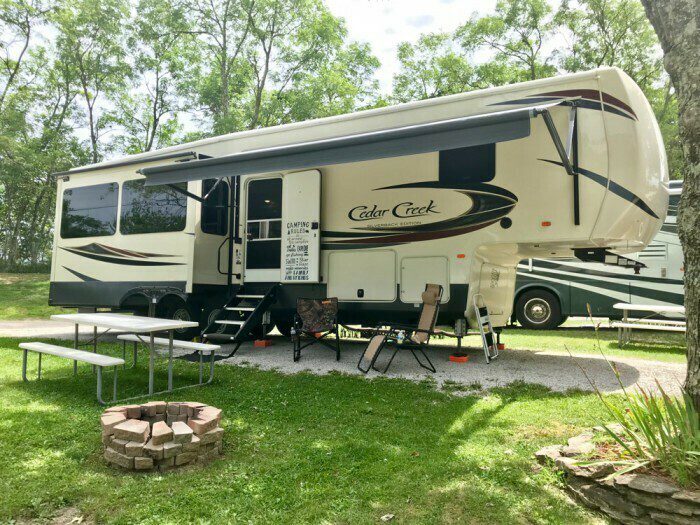 A large Cedar Creek RV is parked on grass beside a picnic table, fire pit, and two lawn chairs, with trees and another RV in the background under a clear sky.