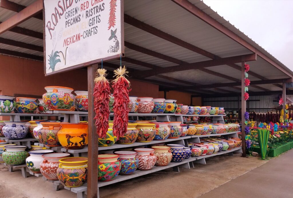 Rows of colorful, hand-painted ceramic pots are displayed on shelves at a roadside shop. A sign advertises Mexican crafts, and dried chili ristras hang near the entrance under a covered roof.