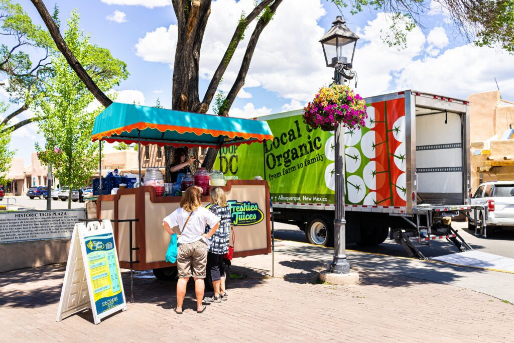 A small juice stand labeled Fresh Juices serves customers on a sunny sidewalk. Behind it, a truck with Local Organic signage and apple graphics is parked. People wait and a hanging flower basket decorates a lamp post.