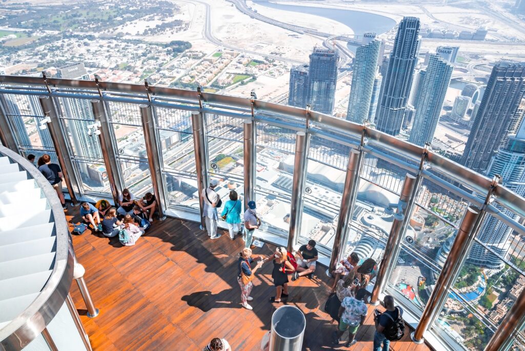 Tourists stand and sit on a wooden deck at a high observation platform with glass walls, overlooking a cityscape with tall skyscrapers, roads, and distant desert.
