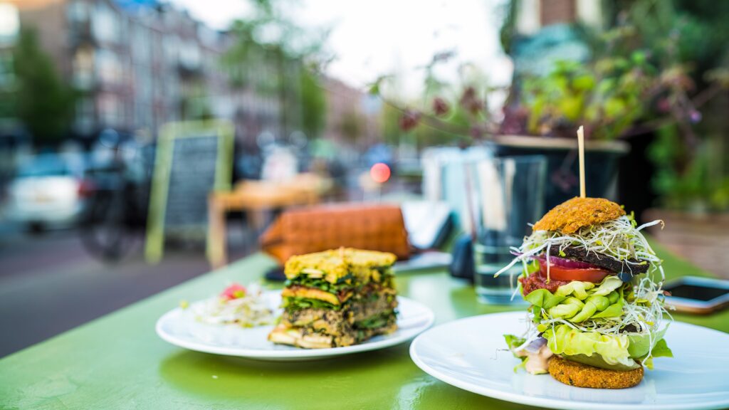 Two plates of food on a green outdoor table; one plate has a tall veggie burger with sprouts and lettuce, the other has a vegetable layered sandwich. The background shows a blurred street scene with buildings and trees.
