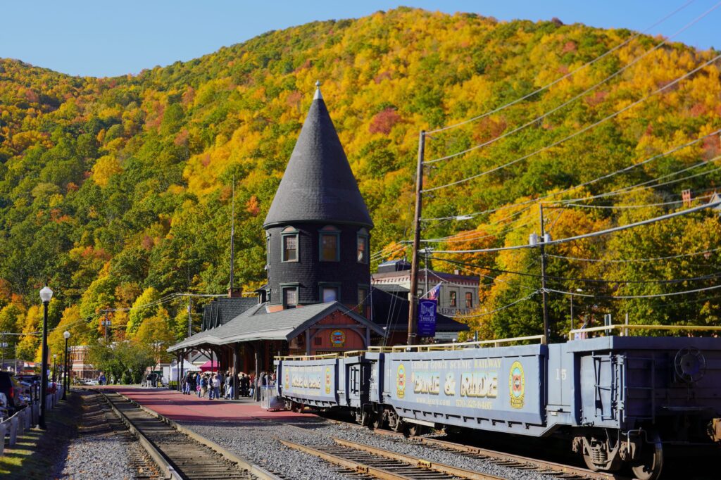 A historic train station with a pointed tower stands beside railroad tracks and freight cars, surrounded by people and vibrant autumn trees covering a hillside in the background.