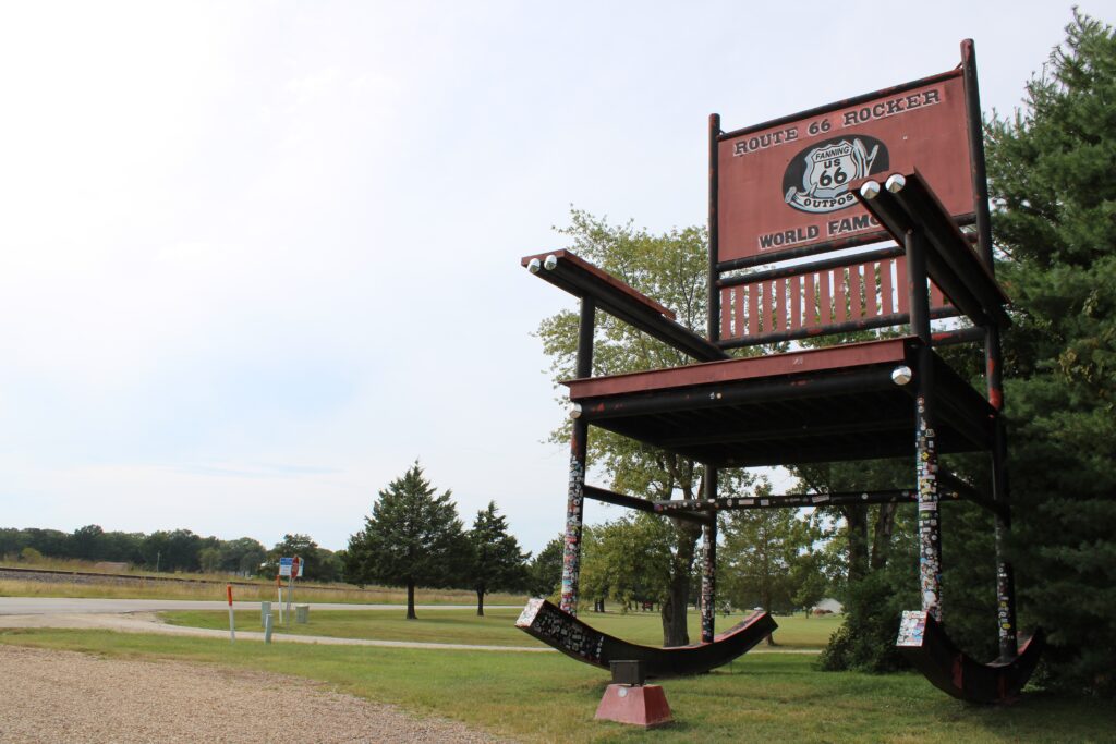 A giant red and black rocking chair with Route 66 Rocker World Famous written on it stands on a grassy area near a road with trees and cloudy sky in the background.