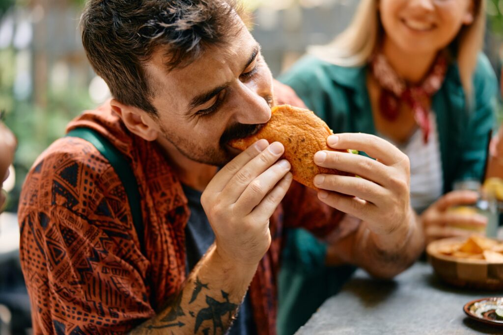 A man with a tattoo on his forearm is eagerly biting into a large sandwich at an outdoor table, with a smiling person sitting beside him and bowls of food in the background.