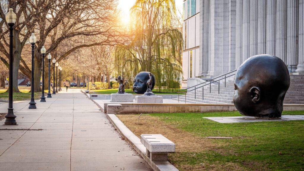 Large bronze sculptures of human heads are displayed outdoors near a grand building with columns. Trees line the sidewalk, and the warm sunlight filters through the branches, creating a serene atmosphere.