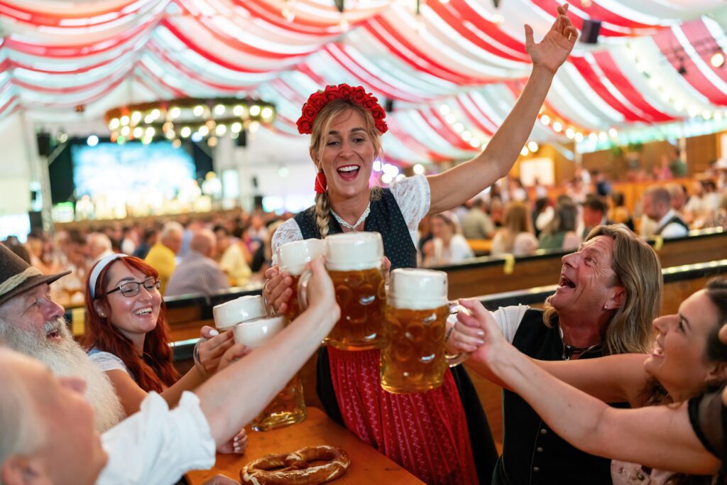 A group of people in traditional Bavarian clothing cheerfully toast with large beer mugs under a festive red and white tent at Oktoberfest, with a pretzel on the table.