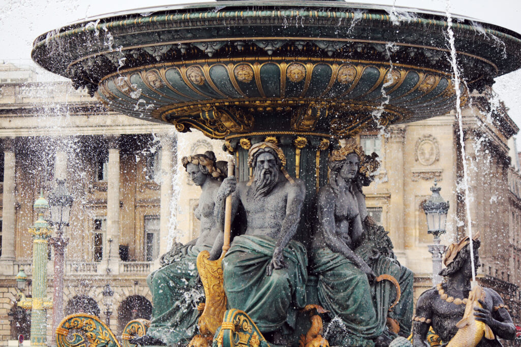 A close-up of a large ornate fountain with detailed statues of seated figures and water spraying, set against the backdrop of a grand historic building.
