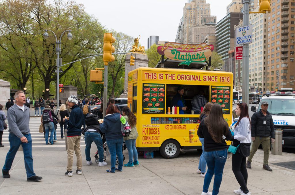 A busy street scene in New York City shows people gathered around a Nathans Famous yellow hot dog stand, with tall buildings and trees in the background and a police vehicle parked nearby.