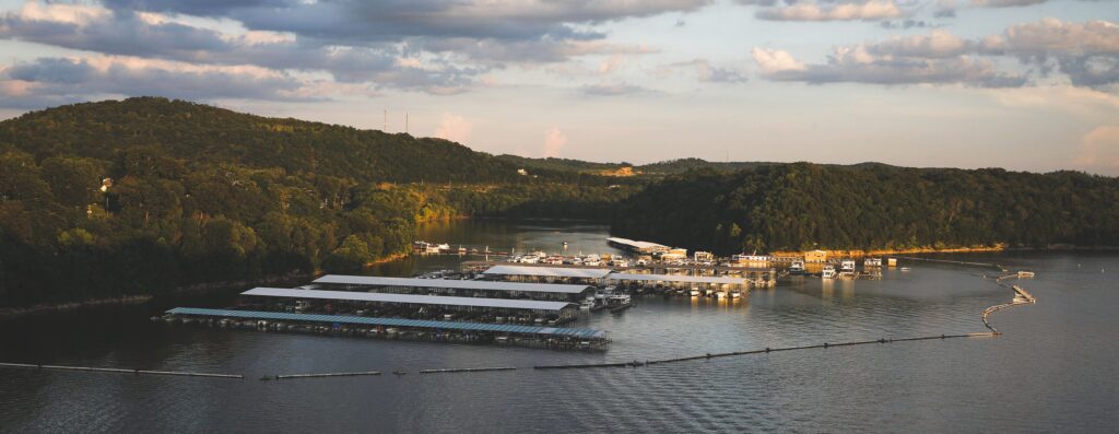 A marina with covered boat docks sits on a calm lake surrounded by green, forested hills under a partly cloudy sky at sunset. Floating barriers extend across the water near the docks.