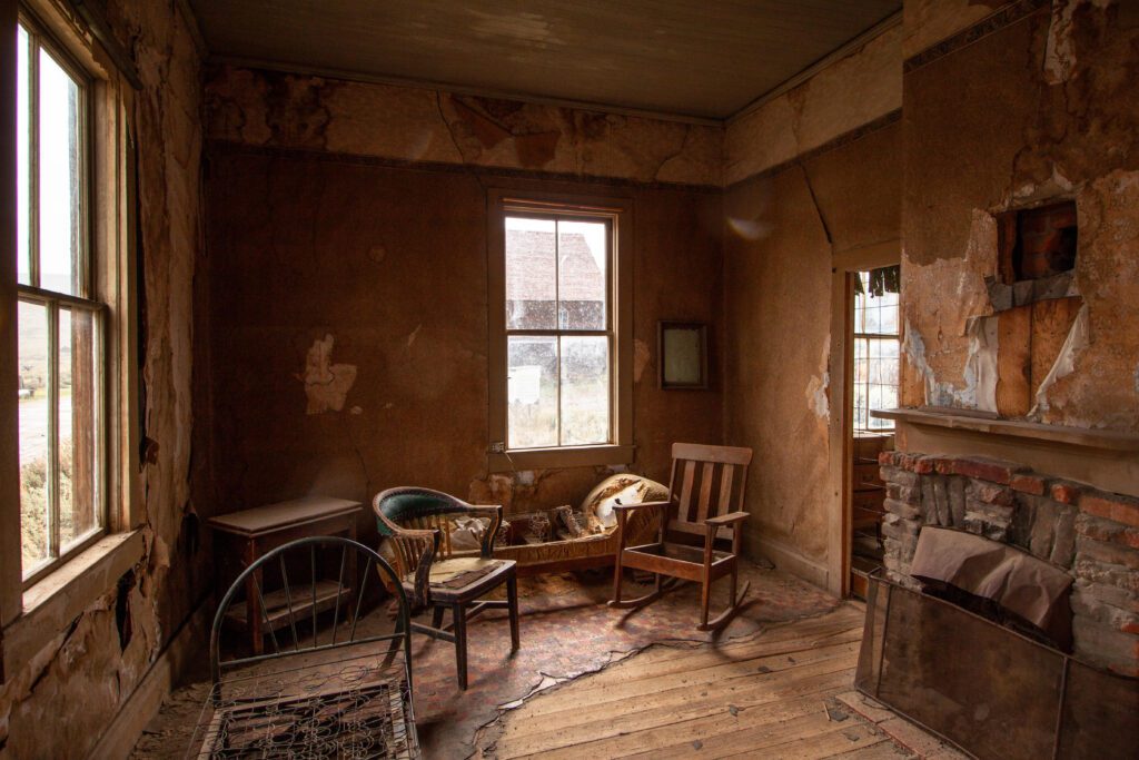 An abandoned, weathered room with peeling walls, old furniture including a crib, rocking chair, and loveseat, wooden floors, a broken fireplace, and sunlight coming through dirty windows.