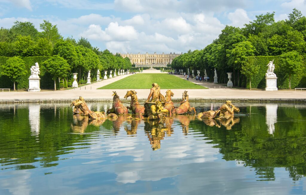 A large palace stands at the end of a manicured garden with symmetrical hedges and statues. In the foreground, a grand fountain with sculpted horses and figures is reflected in a pond under a partly cloudy sky.