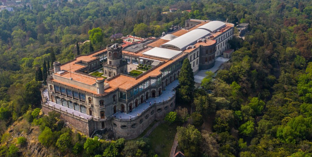 Aerial view of Chapultepec Castle, a large historic building with a central tower and red roofs, surrounded by lush green trees in a forested area.