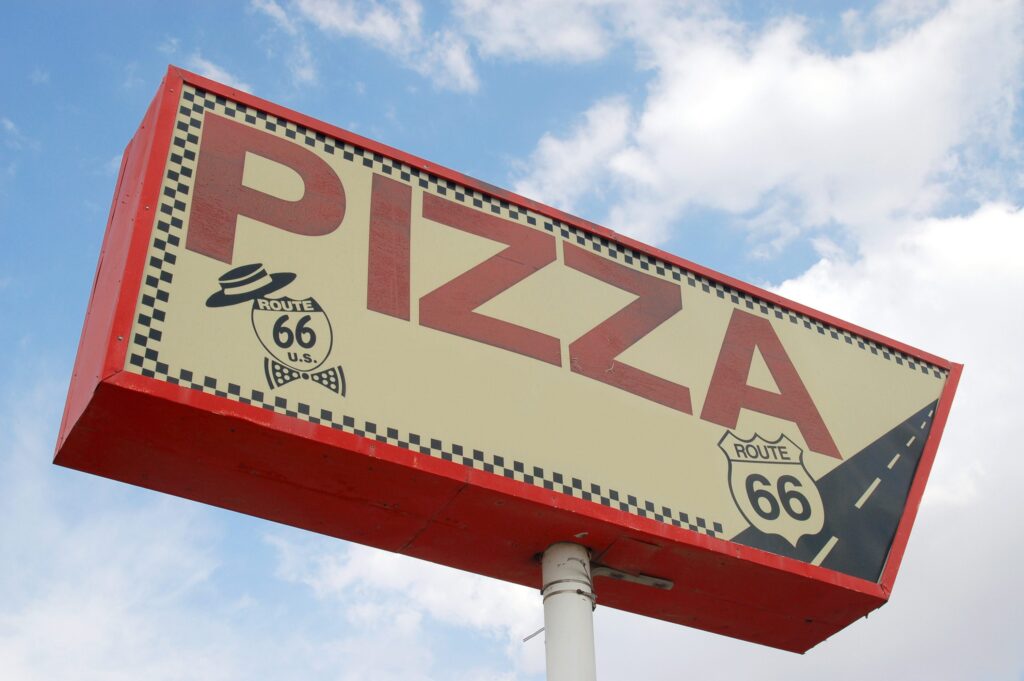 Large retro-style sign with the word PIZZA in bold red letters, displaying two Route 66 highway shields and a checkered border, set against a blue sky with clouds.