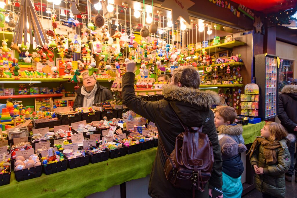 A person points at toys on a brightly lit market stall while children and another adult watch; the stall is filled with colorful toys and small items, creating a lively, festive atmosphere.
