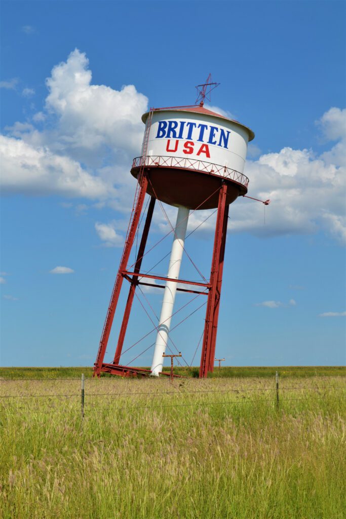 A tilted water tower with BRITTEN USA written on it stands in a grassy field under a blue sky with scattered clouds. The towers red and white structure leans noticeably to one side.