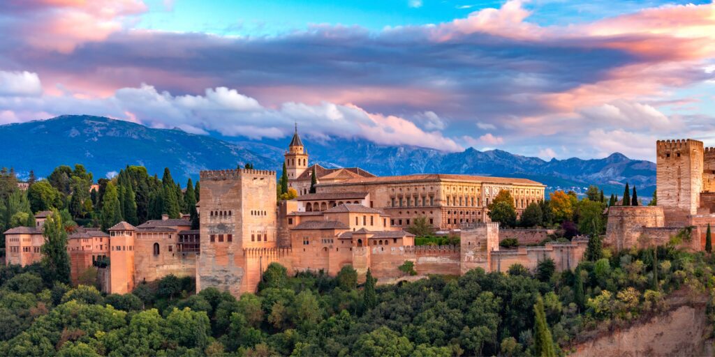 Panoramic view of the Alhambra palace in Granada, Spain, surrounded by green trees with mountains and a colorful, cloudy sky in the background.