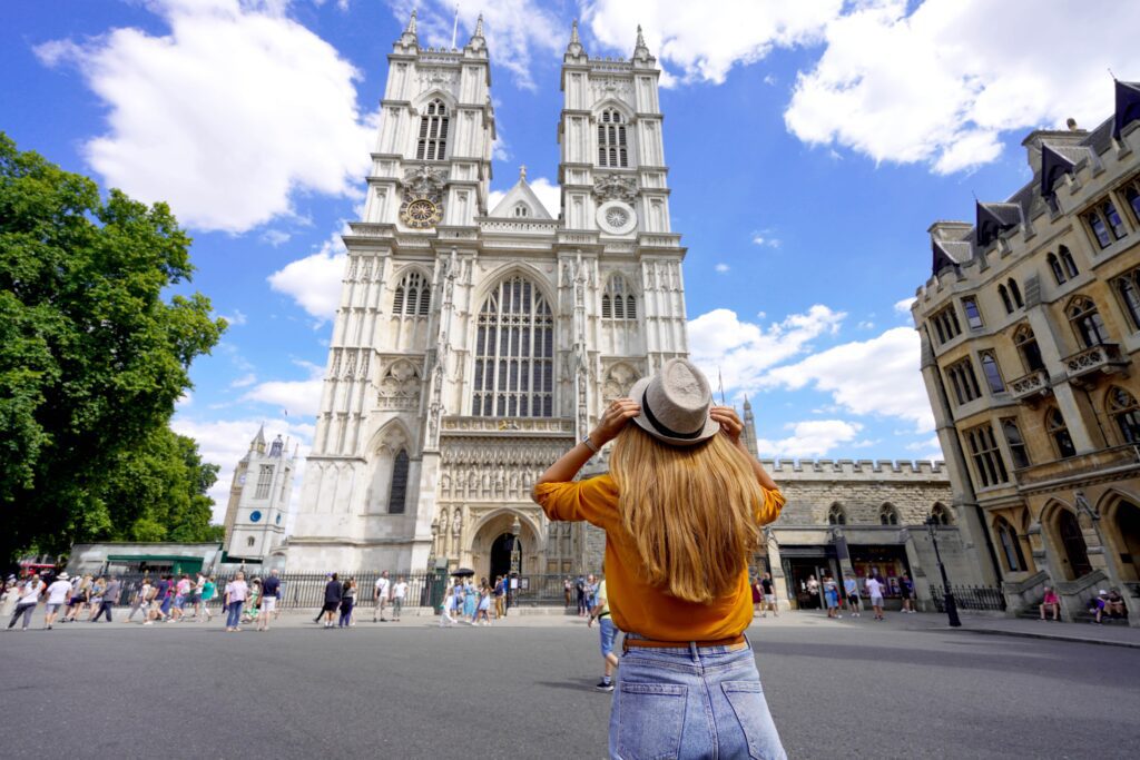 A woman in a yellow shirt and hat stands facing Westminster Abbey in London, with her hands on her hat. Crowds of people walk nearby under a bright blue sky with scattered clouds.