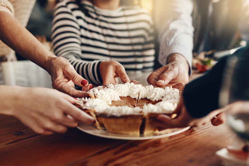 Four people reach in to hold a pie topped with whipped cream, sharing it over a wooden table. A person in a striped shirt sits in the background, out of focus.