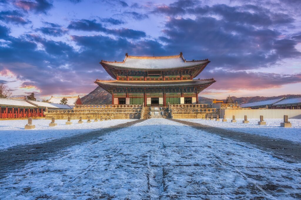 A traditional Korean palace building stands on a snowy courtyard at sunrise, with dramatic clouds and mountains in the background. The detailed architecture is accentuated by the soft morning light.