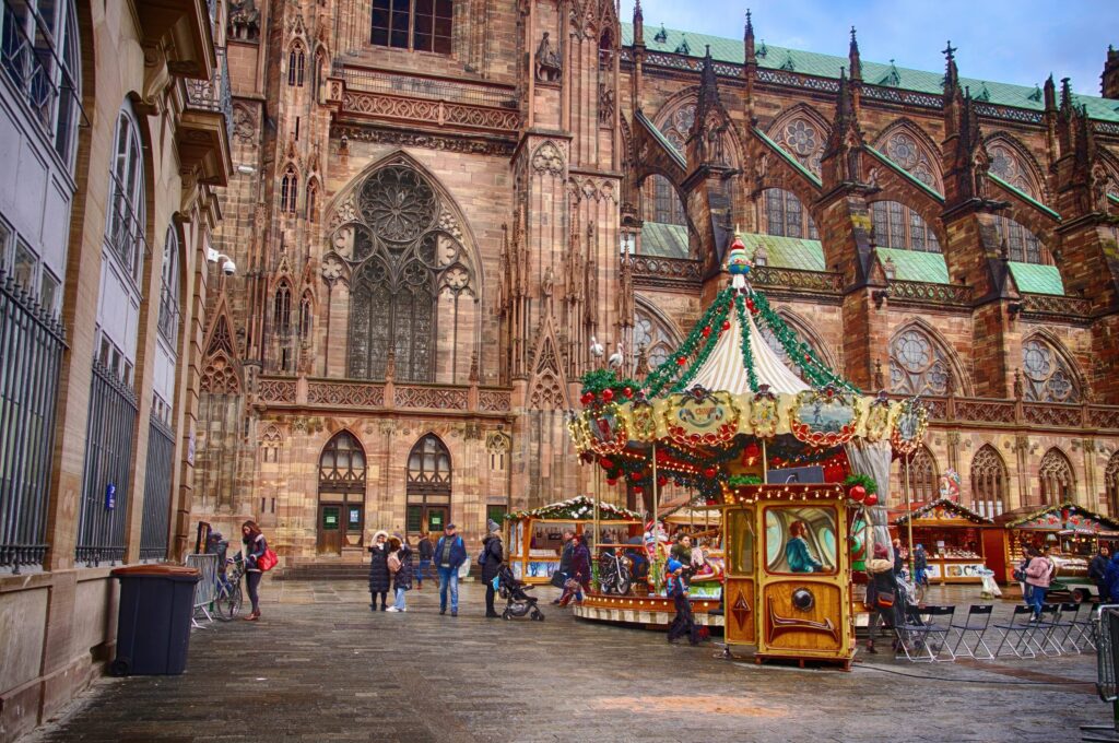 A colorful carousel decorated for Christmas stands in front of a large Gothic cathedral, as people walk around and children ride the carousel. Stalls and festive decorations add to the lively holiday atmosphere.