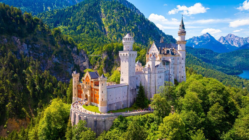 Aerial view of Neuschwanstein Castle in Germany, surrounded by lush green forests and mountains under a blue sky with scattered clouds.