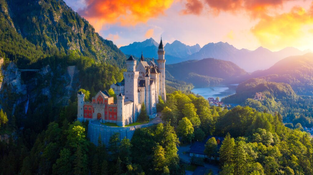 A fairytale-like castle with tall towers sits atop a forested hill, surrounded by mountains and a lake, under a vibrant orange and purple sunset sky.