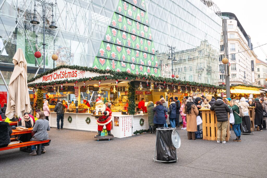 A festive outdoor Christmas market with a decorated stall, a life-size Santa statue, and crowds of people gathering. A large modern glass building is in the background, adorned with a giant Christmas tree pattern.