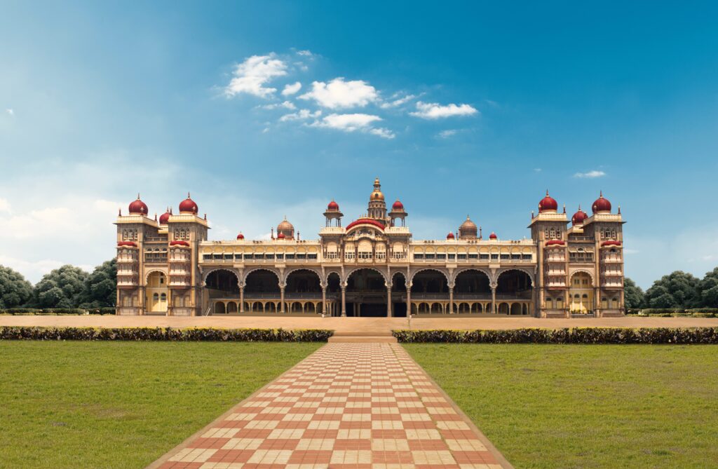 A grand historic palace with red domes and ornate details stands under a bright blue sky, viewed from a tiled pathway across a large green lawn.