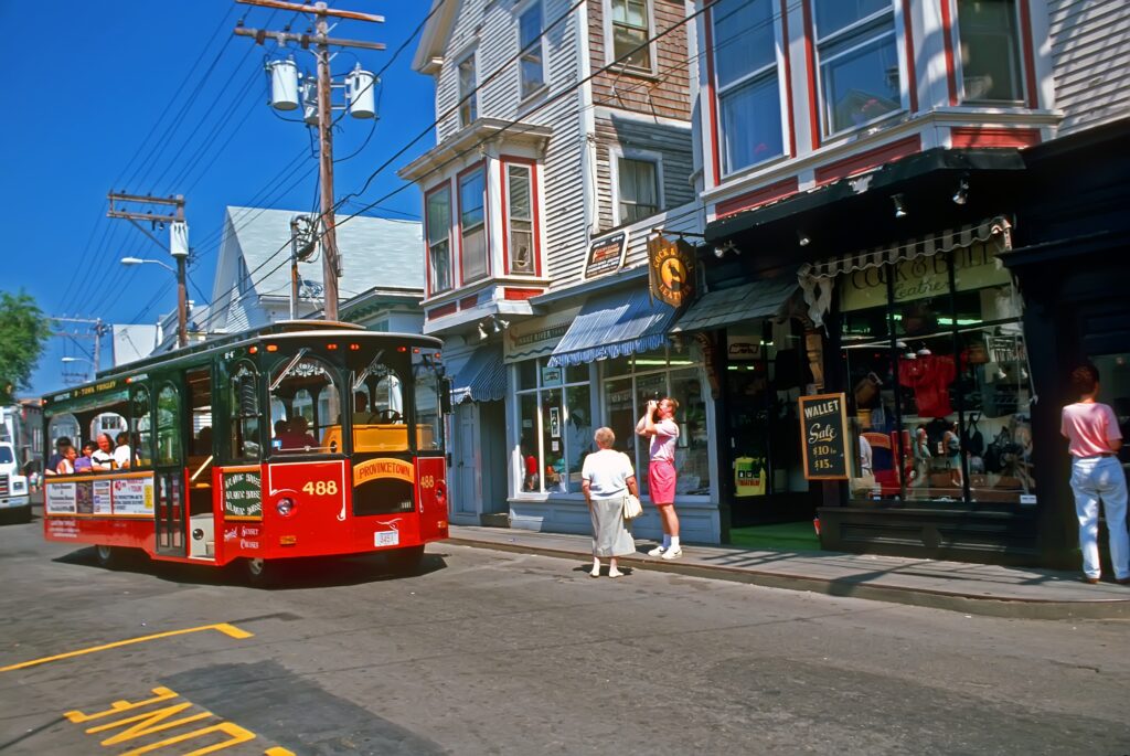 A red trolley bus drives down a street lined with colorful shops and wooden buildings. People stand on the sidewalk, some window shopping and others taking photos. The scene is bright and lively on a sunny day.