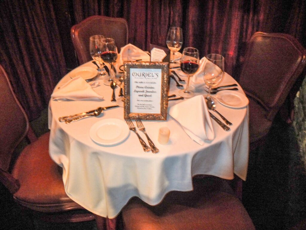A round dinner table for four set with white linens, plates, silverware, wine glasses with red wine, and folded napkins. A framed menu stands in the center of the table. Four brown chairs surround the table.