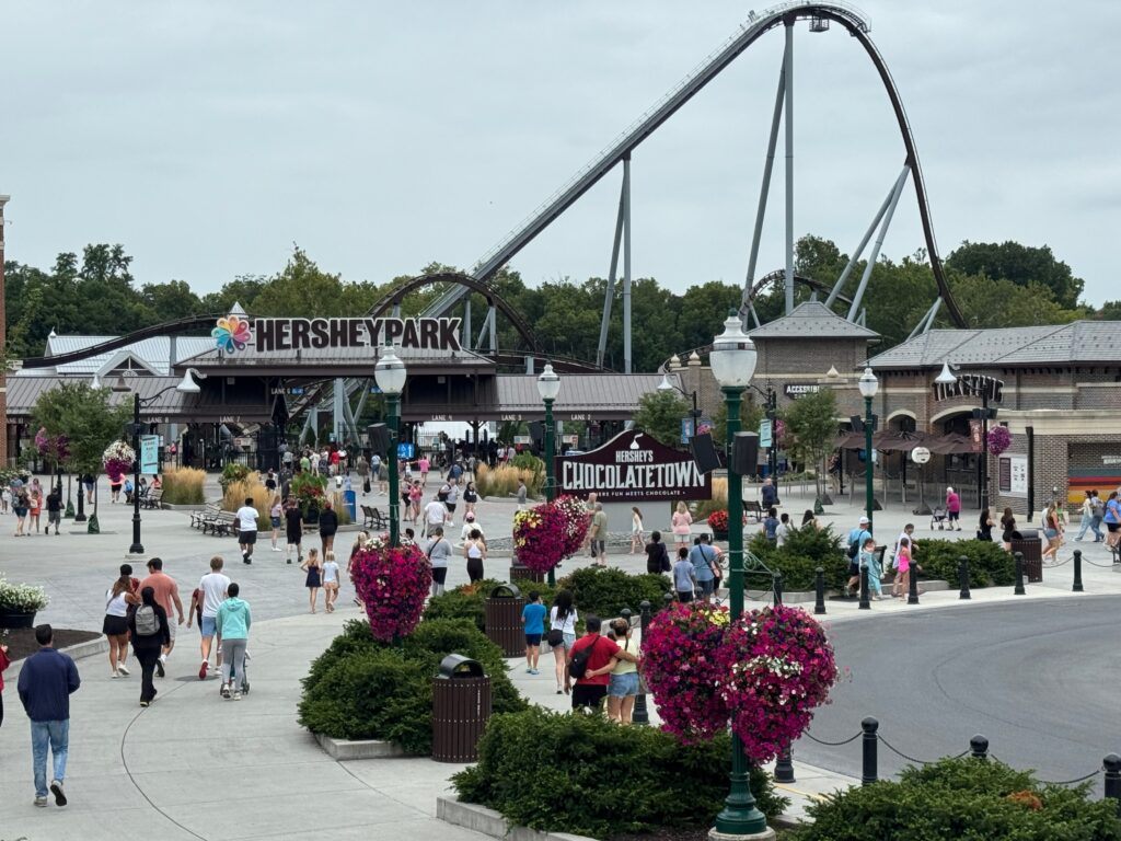 People walk around Hersheypark entrance and Chocolatetown area, with a large roller coaster in the background, flower planters lining the walkway, and overcast sky above.