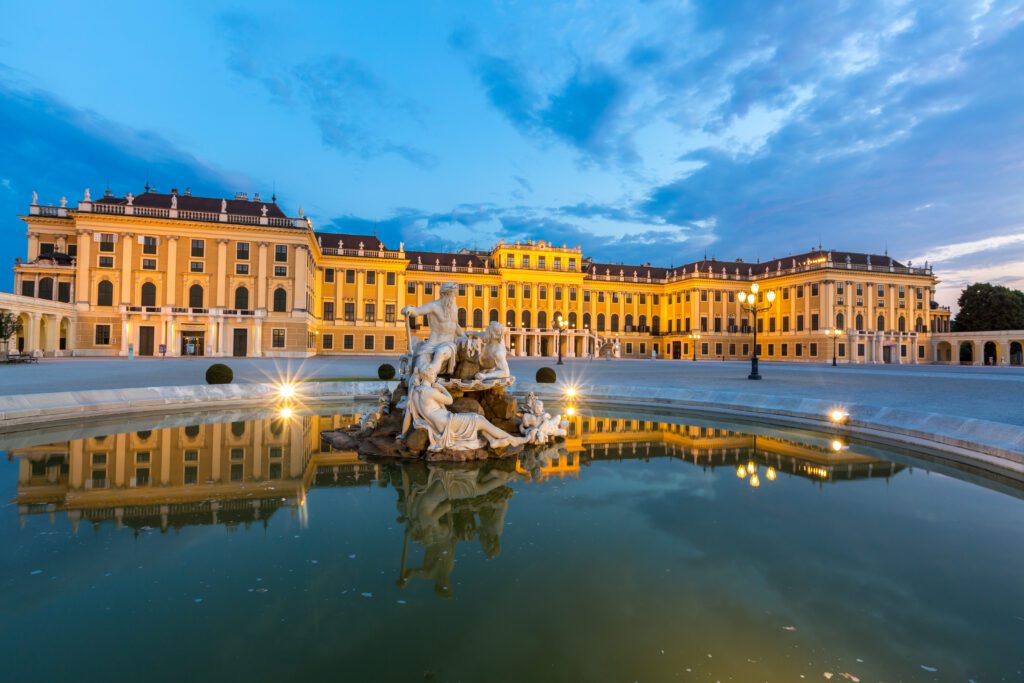 The illuminated Schönbrunn Palace in Vienna at dusk, with a baroque fountain and statues reflected in the still water in the foreground.