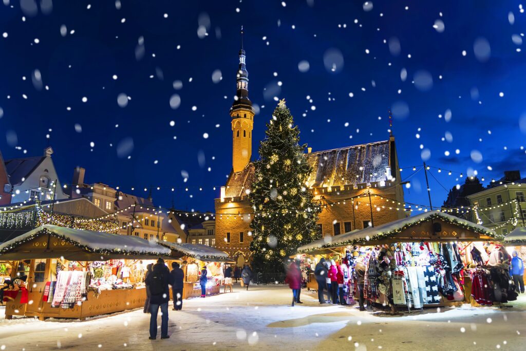 A festive Christmas market at night with wooden stalls, a large decorated Christmas tree, people shopping, and snow falling. Warm lights illuminate the scene and a tall, lit-up church tower stands in the background.