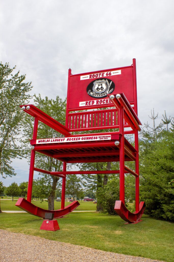 A giant red rocking chair labeled Route 66 Red Rocker stands on a grassy area near trees, with a sign reading Worlds Largest Rocker - Guinness. The sky is overcast.