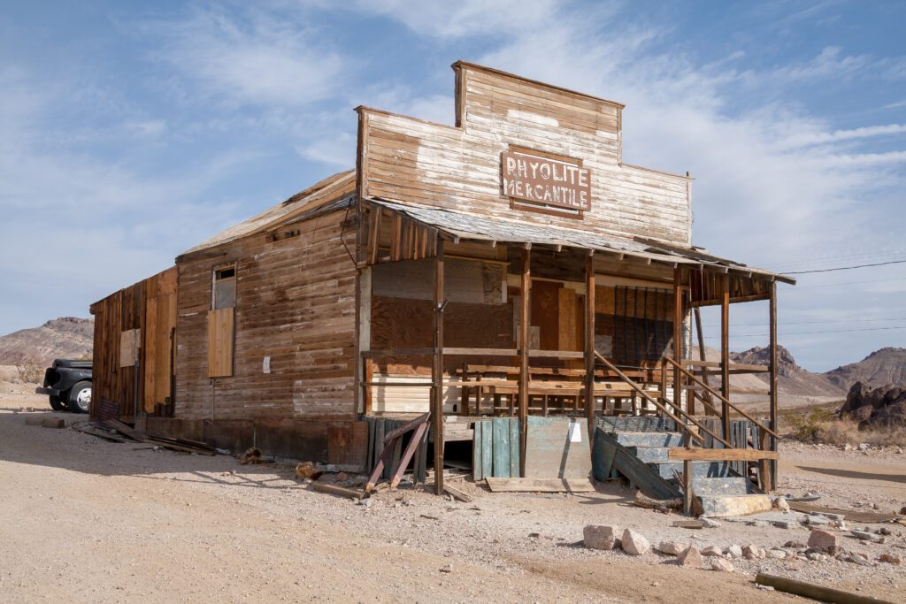 A weathered, abandoned wooden building labeled “Rhyolite Mercantile,” with peeling paint and broken steps, sits in a desert landscape under a partly cloudy sky. Rocky hills and a parked vehicle are visible in the background.