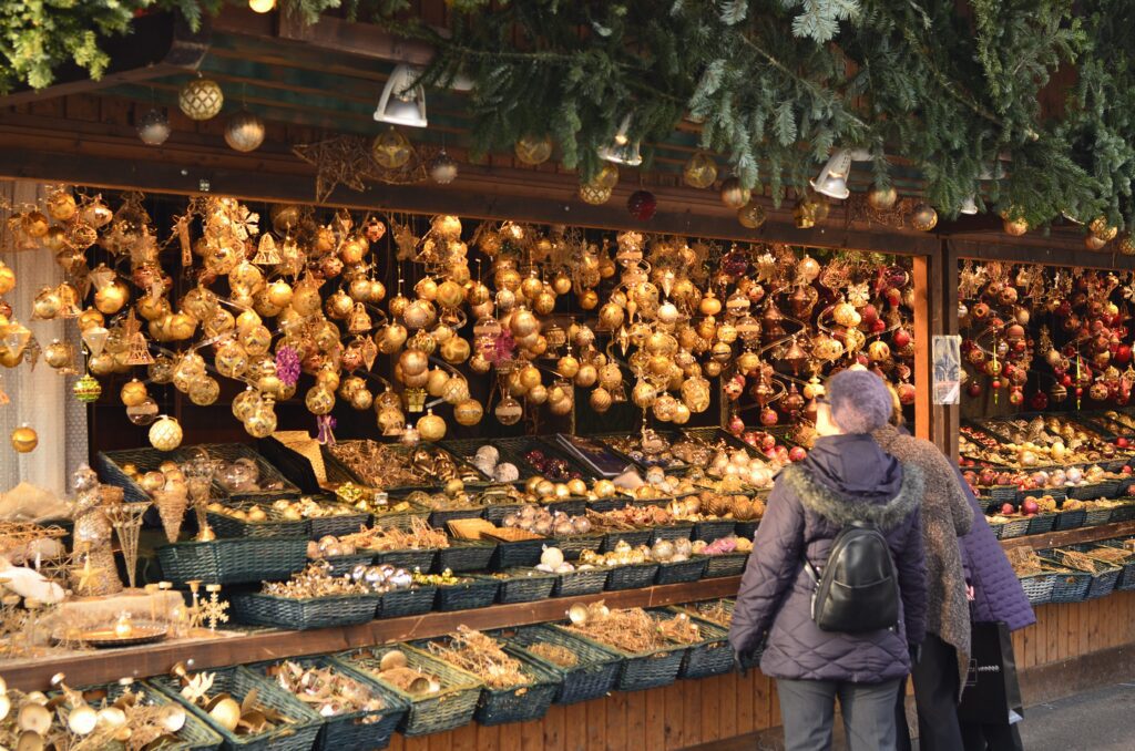 Two people browse a festive market stall decorated with hanging golden ornaments and filled with baskets of holiday decorations. Green pine branches hang overhead, adding to the seasonal atmosphere.